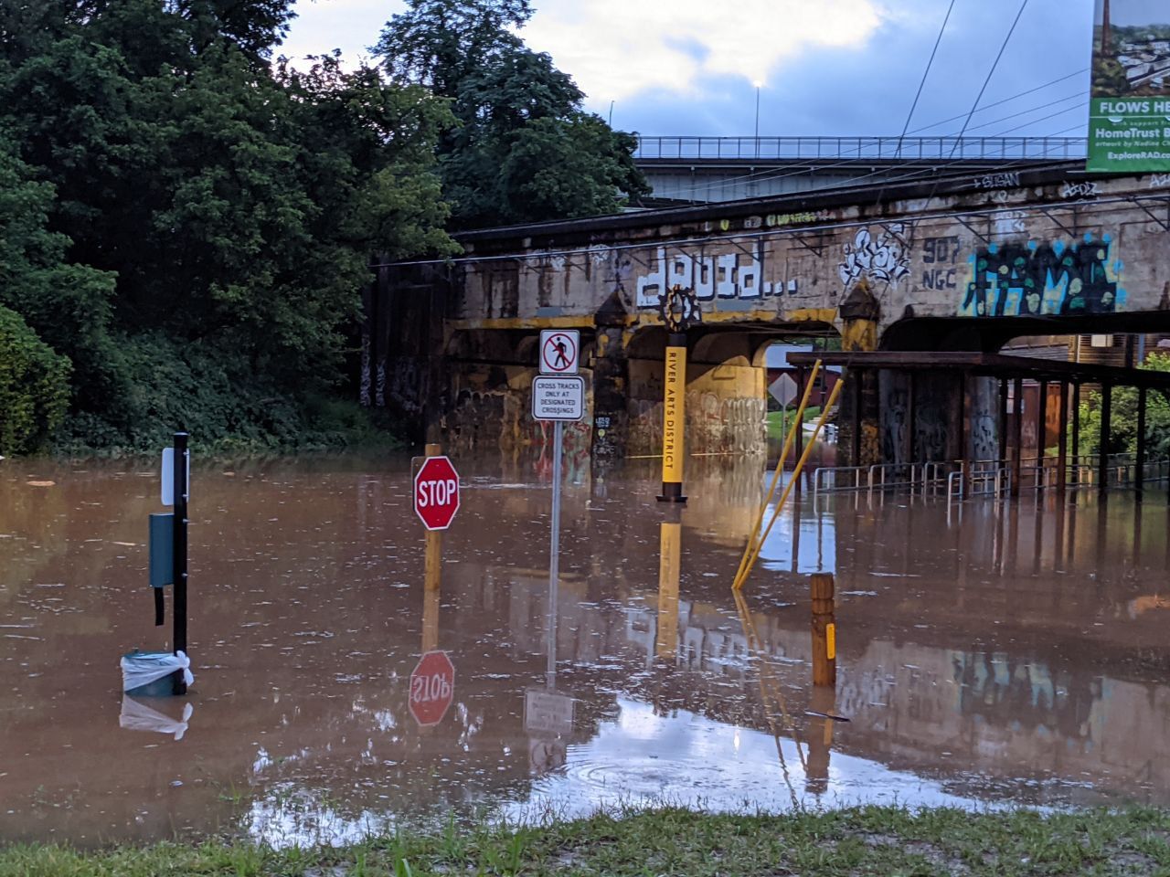 Flooded street with railroad bridge over top. Stop sign and public trashcan peek over top of the water. Bridge has a sign that reads “River Arts District”