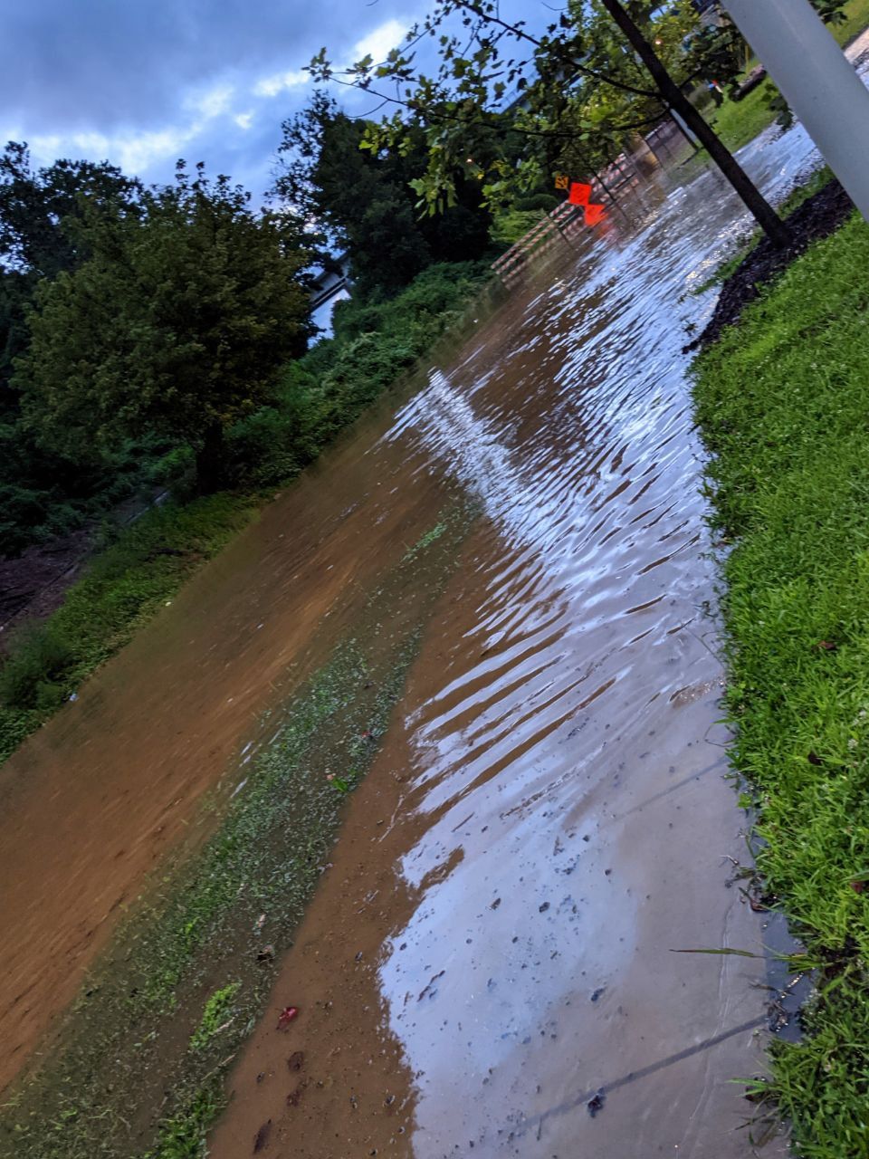 Sidewalk and road in river arts district covered in flood waters. Orange “Road closed” sign in the background with red and white striped barricade