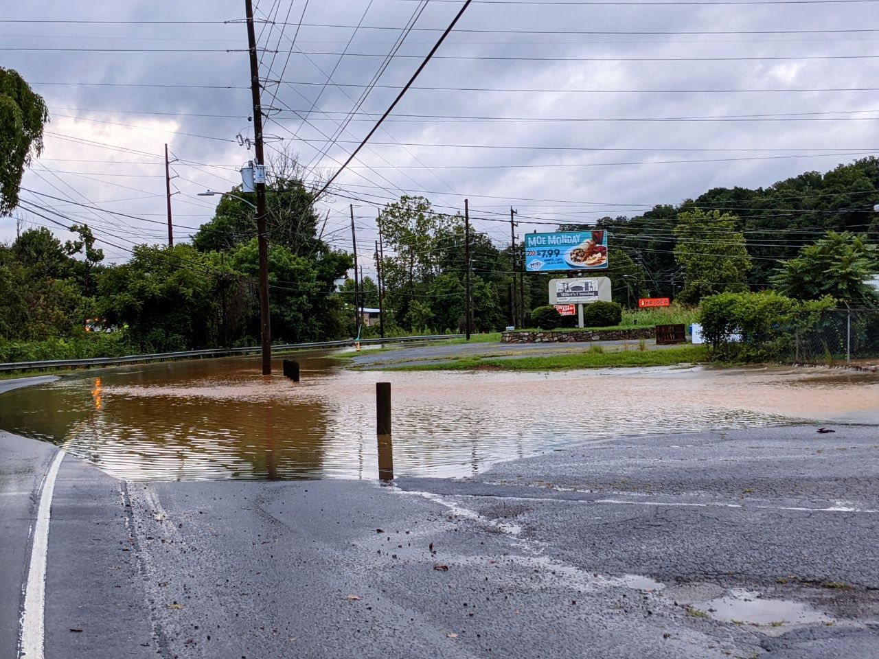 Road bends around a curve, water is flowing over the road and a parking lot beside the road. A billboard in the background reads “Moe Monday” with a burrito advertisement