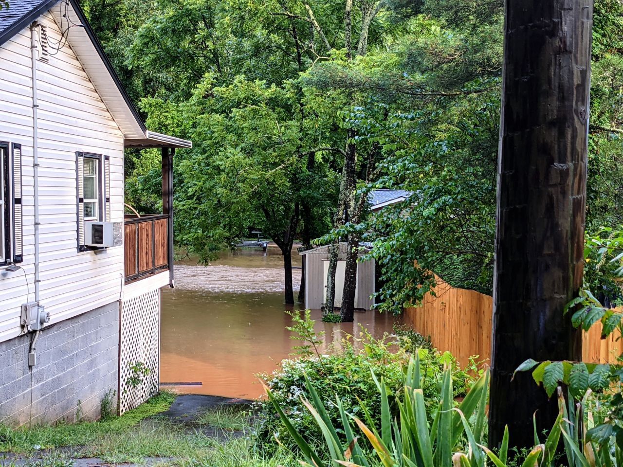 White house on left side of frame, the house’s backyard is flooded.Sheds and other houses are submerged in the water.