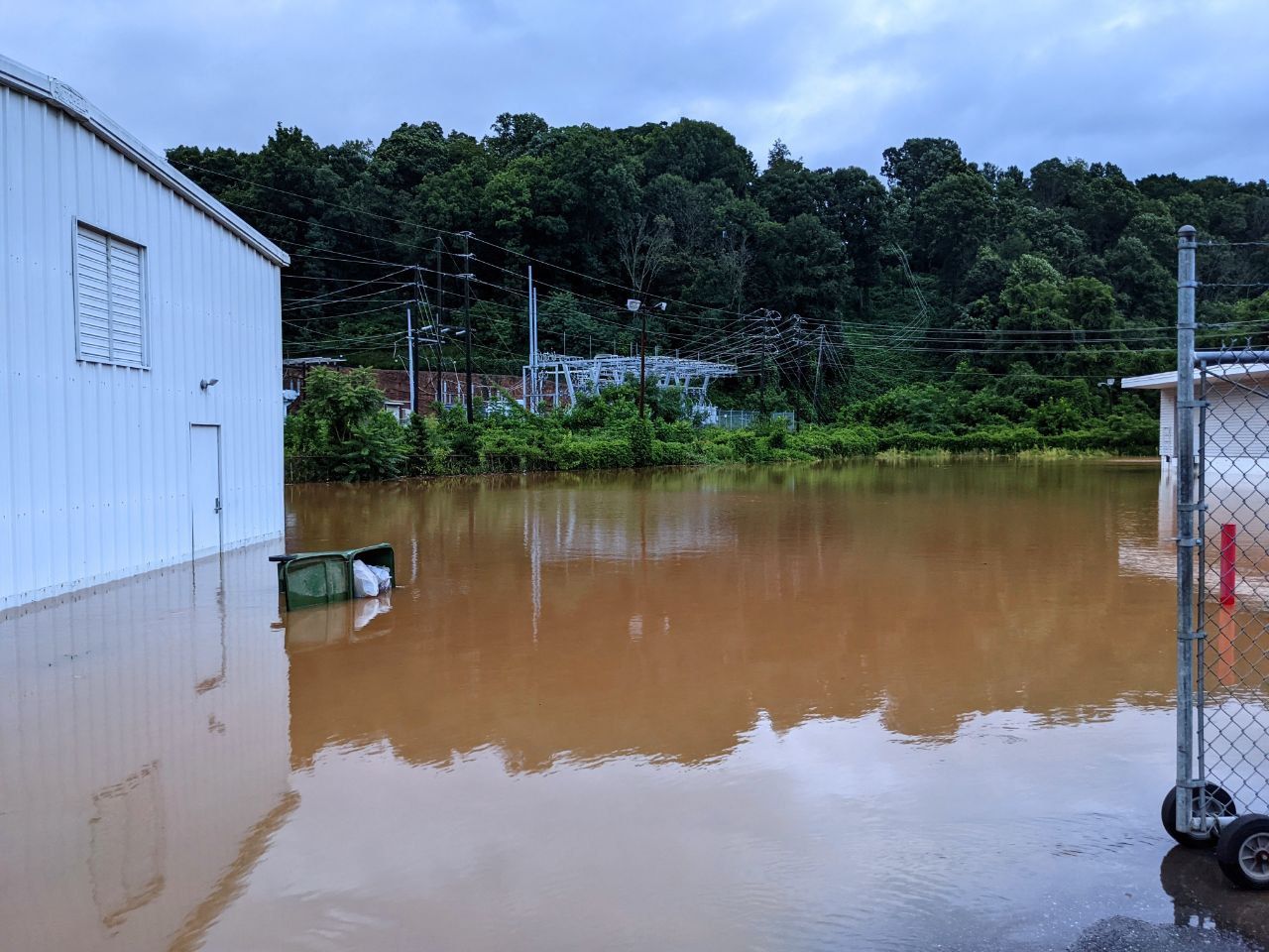 Brown water filling a parking lot, two warehouse style buildings visible, along with an overturned green city trashcan