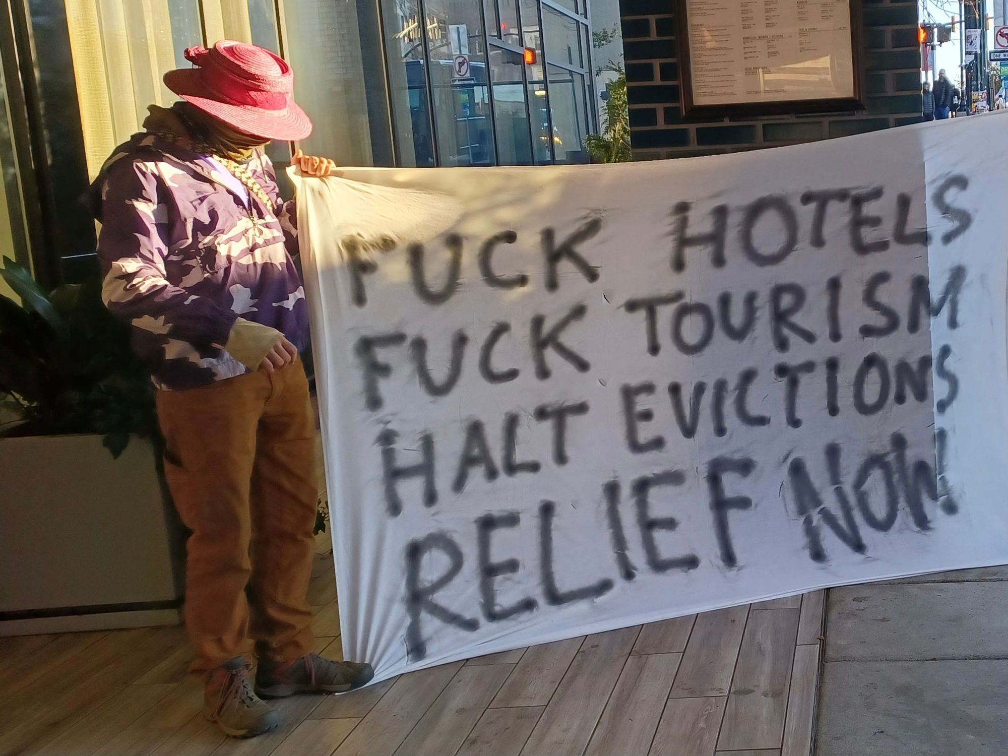 One person, with their face obscured by a broad sun hat, standing in front of a hotel holding a banner that reads reads "Fuck Hotels, Fuck Tourism, Halt Evictions, Relief Now"