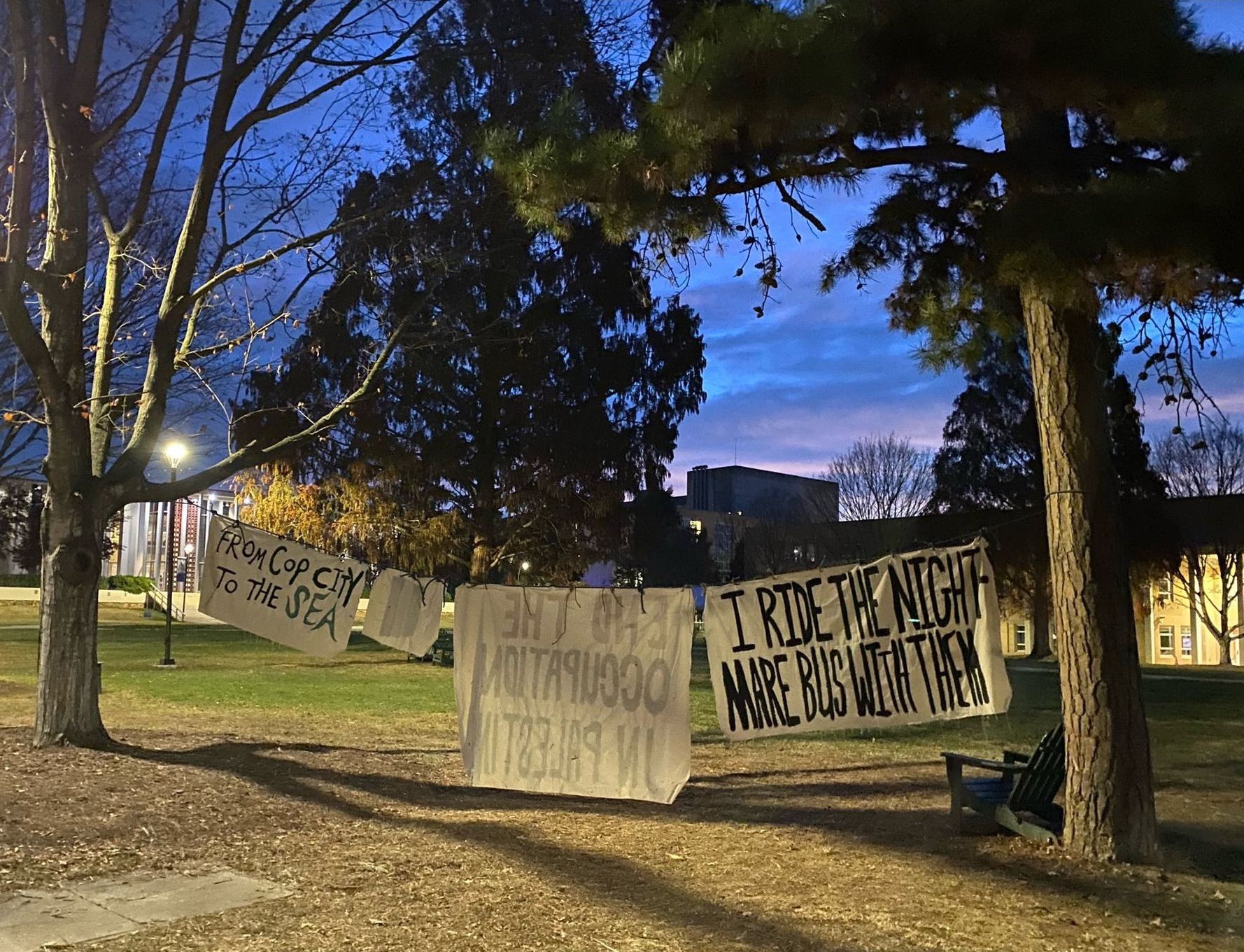 Four painted white banners of various sizes hang from a rope between two trees in front of Karpen Hall on the UNC Asheville campus. The banners facing the viewer read, "From Cop City To The Sea" and "I Ride The Nightmare Bus With Them."".