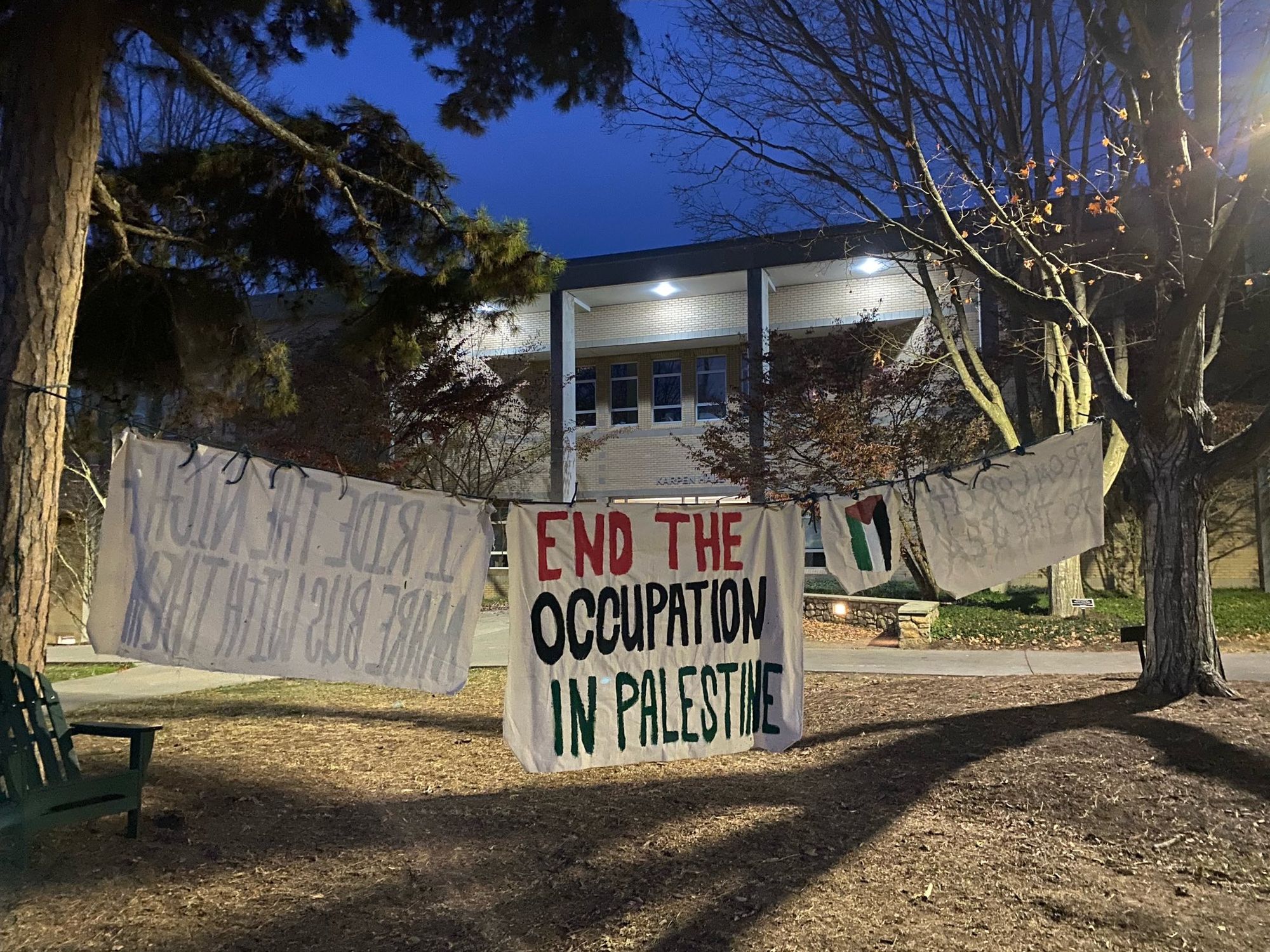 Four painted white banners of various sizes hang from a rope between two trees in front of Karpen Hall on the UNC Asheville campus. The banner facing the viewer reads, "End the Occupation in Palestine".